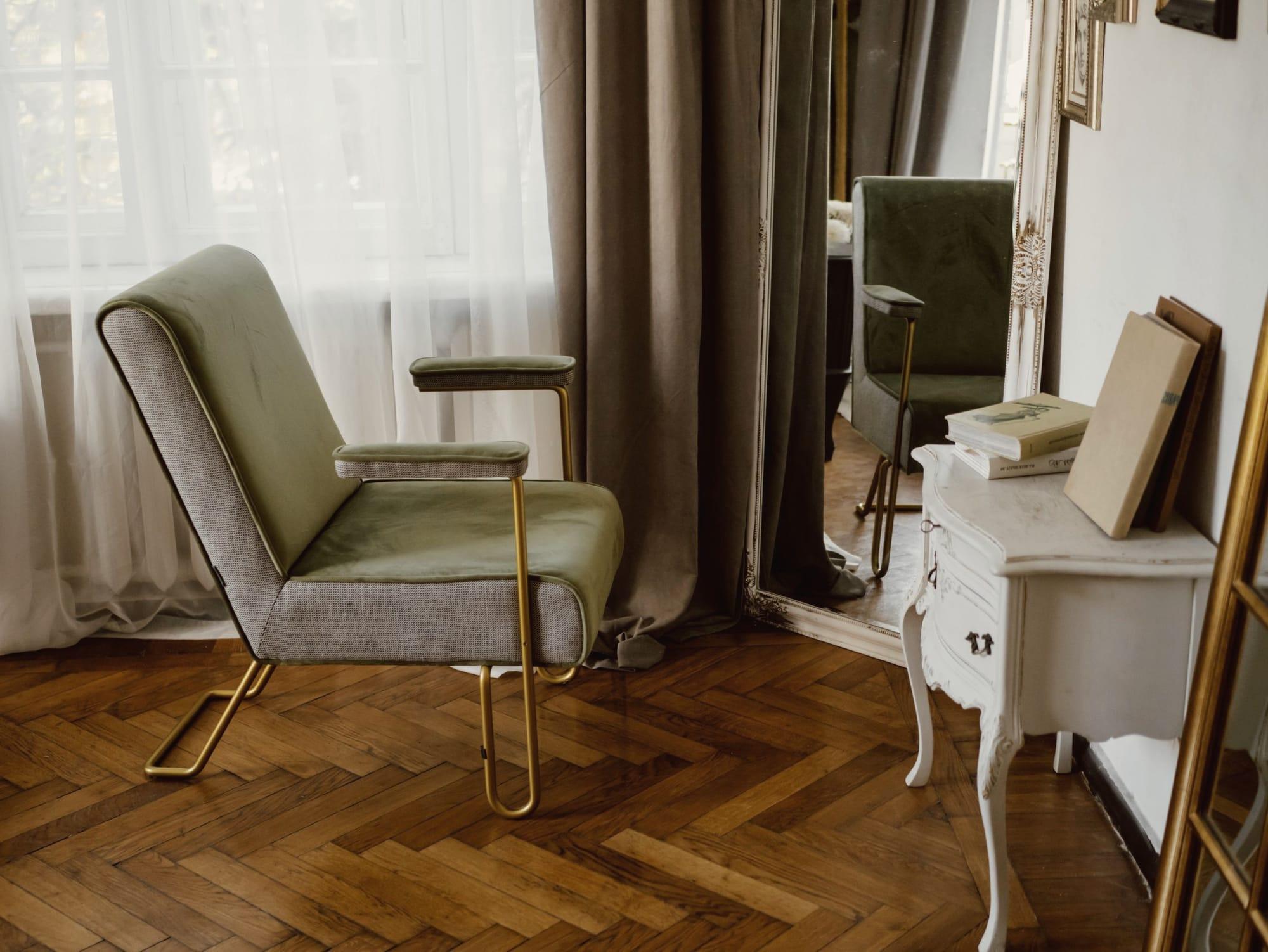 Un salottino con una poltrona in velluto verde salvia, uno specchio dalla cornice intagliata, tende color tortora e parquet a spina di pesce